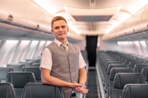 Flight attendant leaning up against seat smiling. He's wearing a white short sleeve dress shirt with a grey vest.